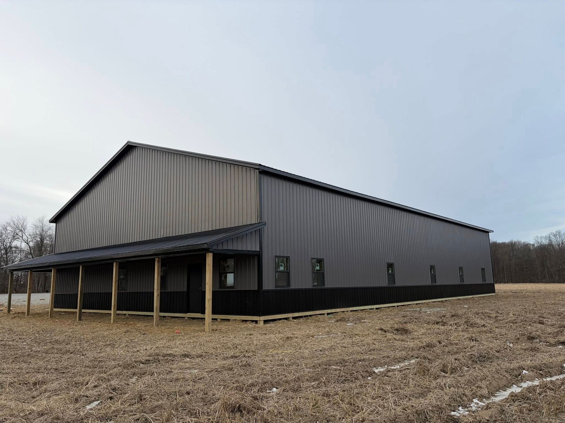 Large gray metal barn-like building with a porch, standing in a muddy field under an overcast sky