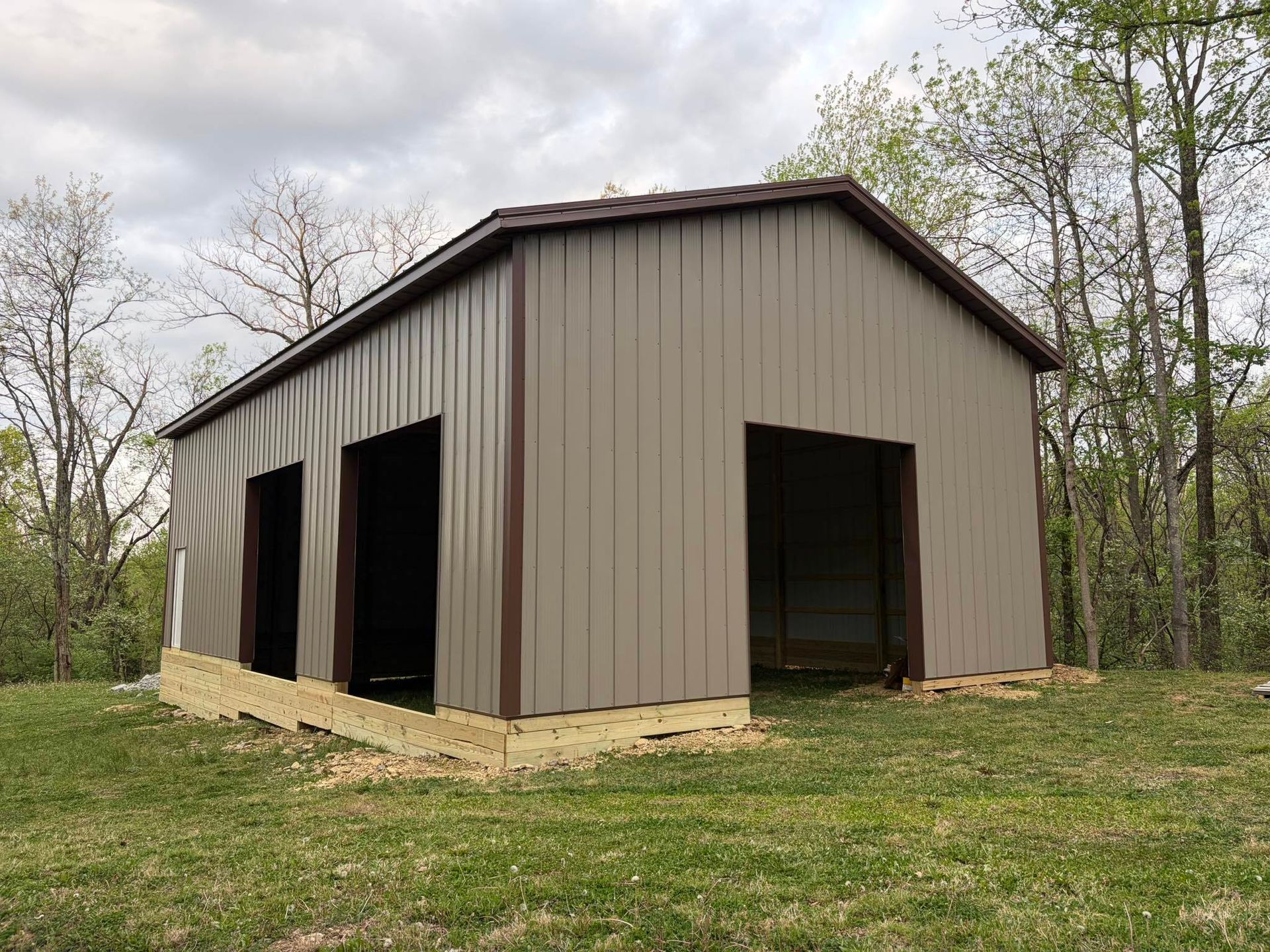 Brown metal shed with large open bays on a grassy lot surrounded by trees