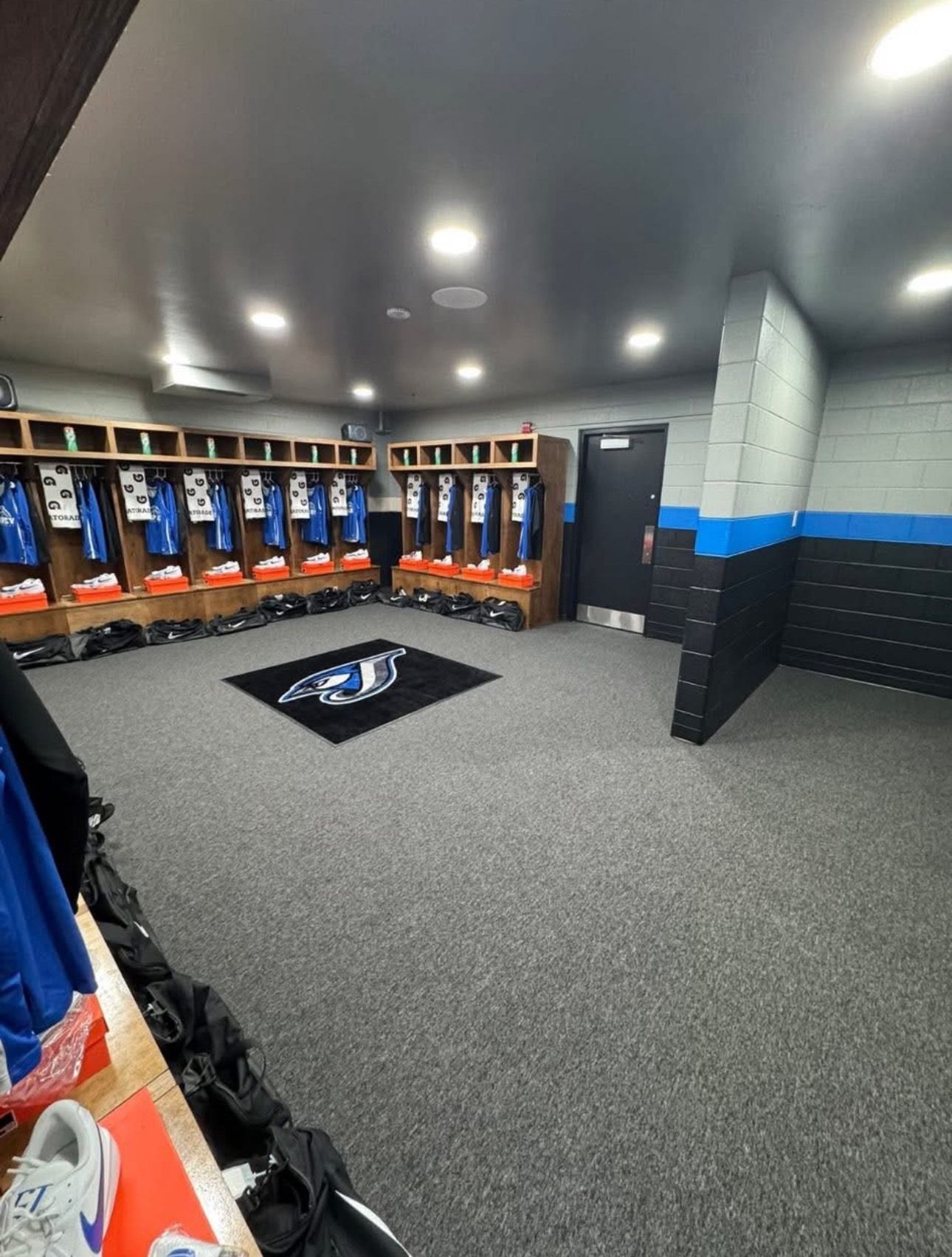 Empty sports locker room with blue-and-black lockers, gray carpet, and a ceiling-mounted TV.