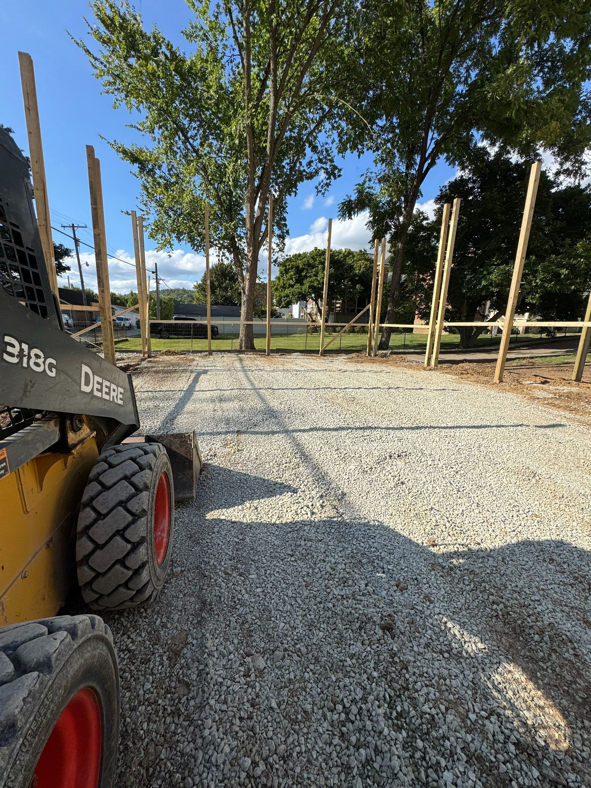 Construction site with gravel ground, trees, and a skid steer on the left.