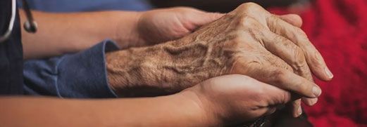 Hands gently holding an elderly person's wrinkled hand on a chair