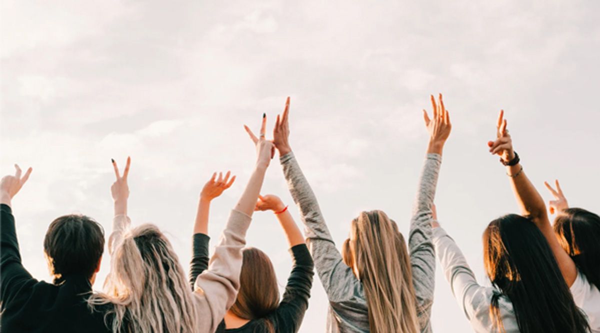Back view of a crowd with raised hands at a concert, wearing light jackets, against a bright sky