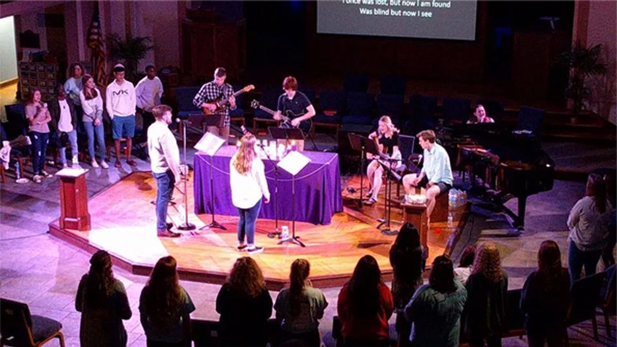 Church worship band performing on stage with singers, musicians, and audience standing in front