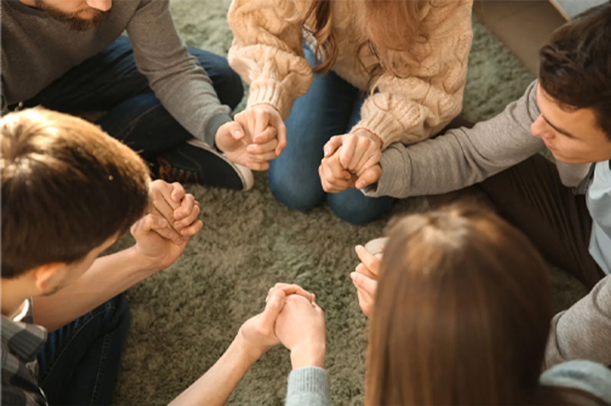 People seated in a circle holding hands, suggesting a group support or prayer meeting.