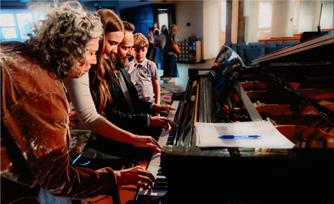 People gathered around a piano, watching and playing keys in a bright indoor hall.
