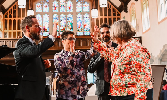 Four people in a church aisle clinking glasses during a toast, with stained glass windows behind them