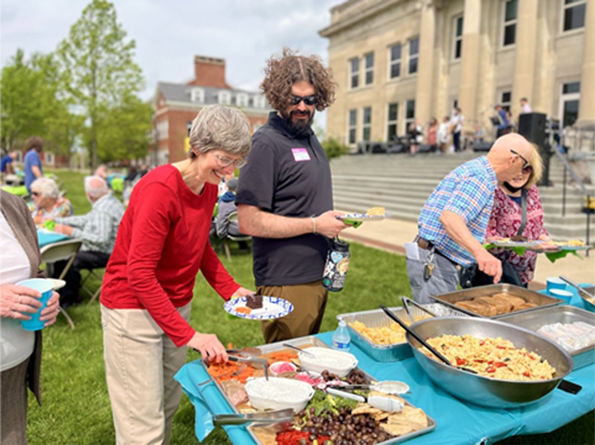 People serving food at an outdoor buffet table on a lawn near a building