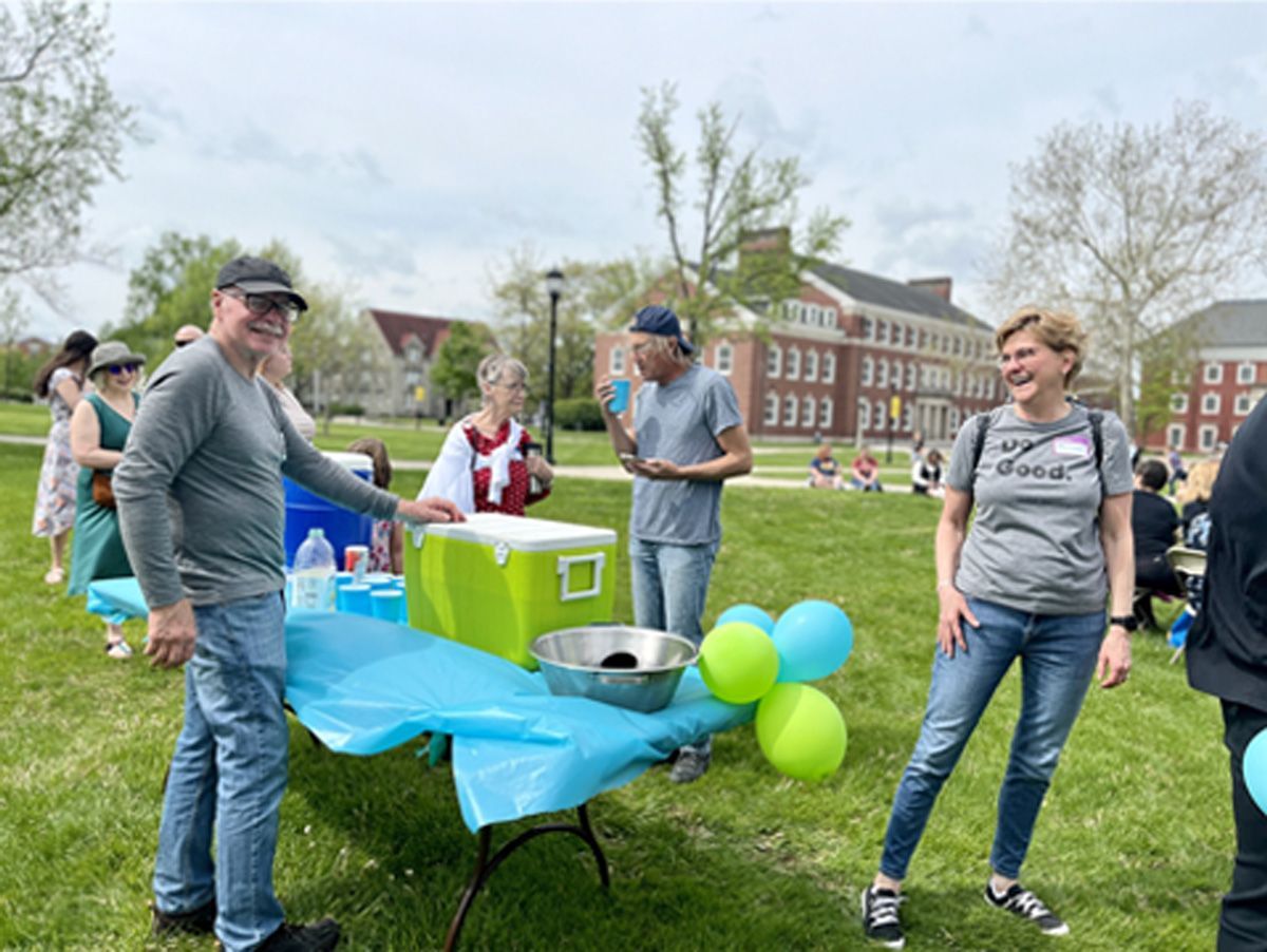 People at an outdoor event around a blue table with green boxes and balloons on a grassy campus.