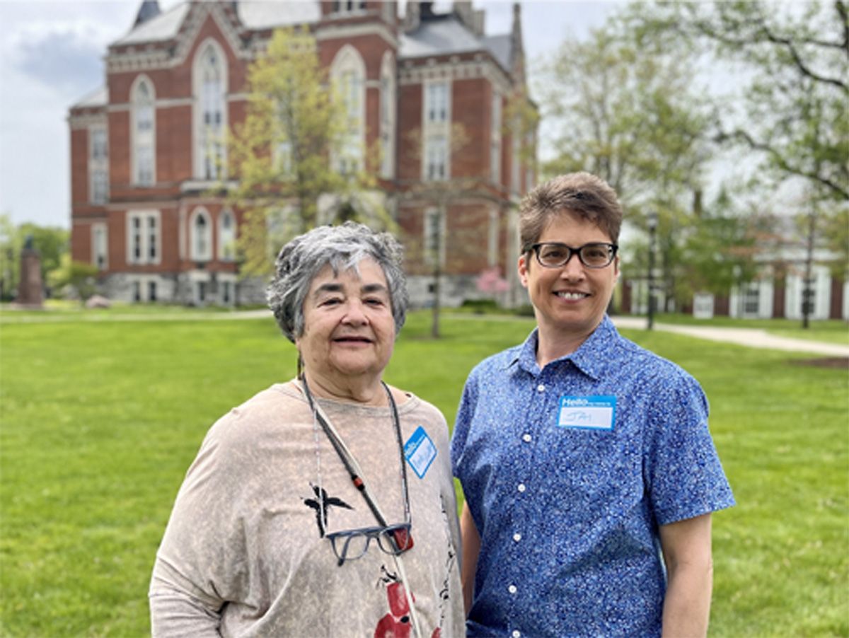 Two people standing on a lawn in front of a brick building, smiling at the camera.
