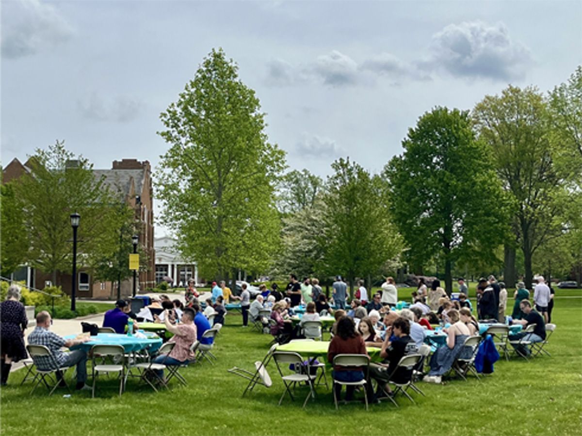 People seated at tables on a grassy lawn in a park, with trees and buildings in the background.
