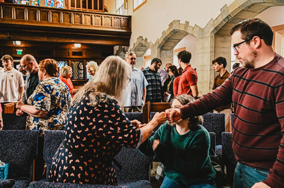 People mingling in a hall, with one man helping another seated person near a table.