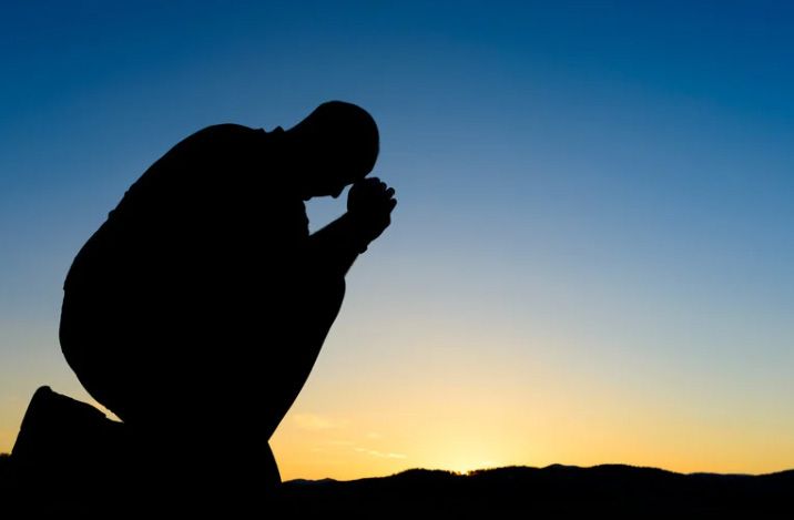 Silhouette of a person kneeling in prayer at sunset against a blue-orange sky