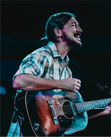 Singer-guitarist performing onstage under blue lighting, smiling while playing an acoustic guitar