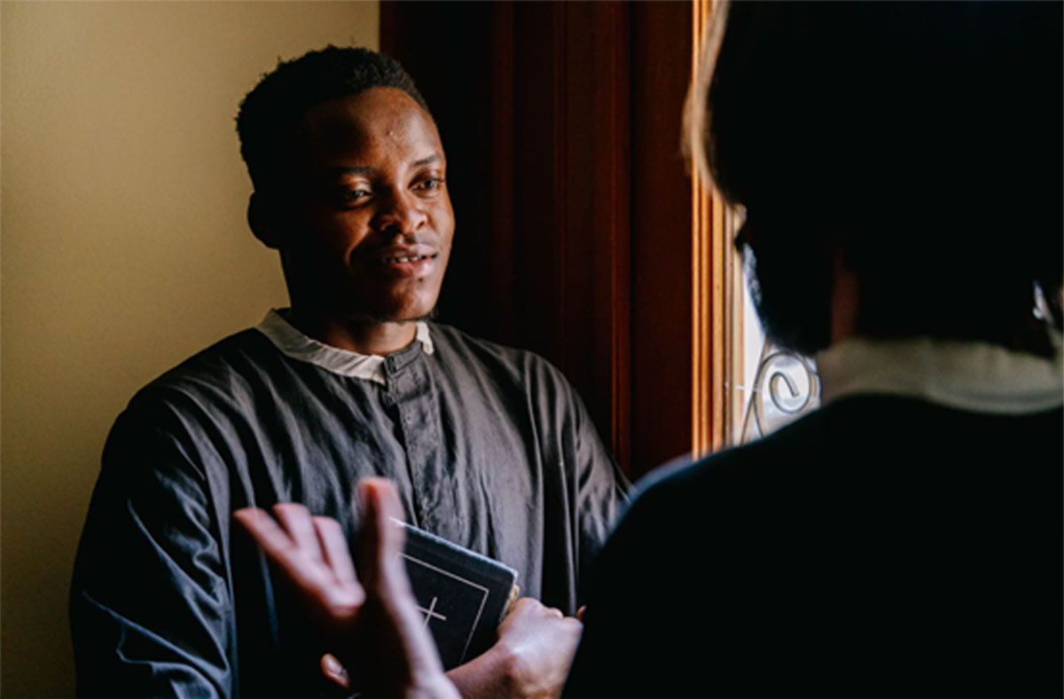 Two people talking indoors by a window, one holding a book and gesturing with a raised hand.