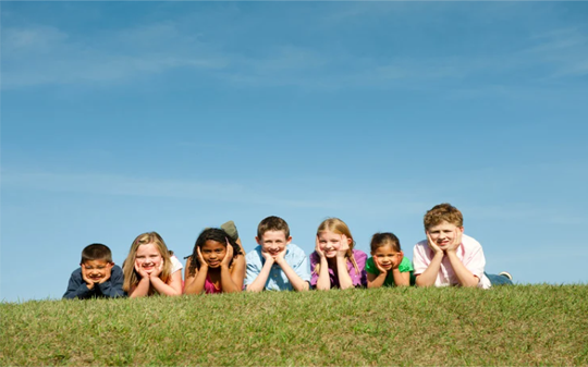 Seven children lying in grass under a blue sky, smiling with hands under their chins