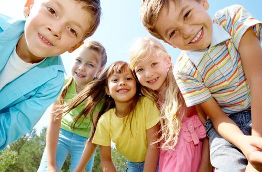 Five smiling children lean close together outdoors in bright sunlight, wearing colorful casual clothes.