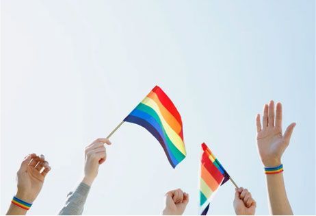 Hands waving rainbow pride flags against a bright sky