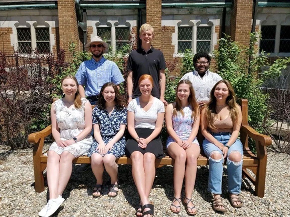 Seven people posing on benches in a garden courtyard in front of a brick building.