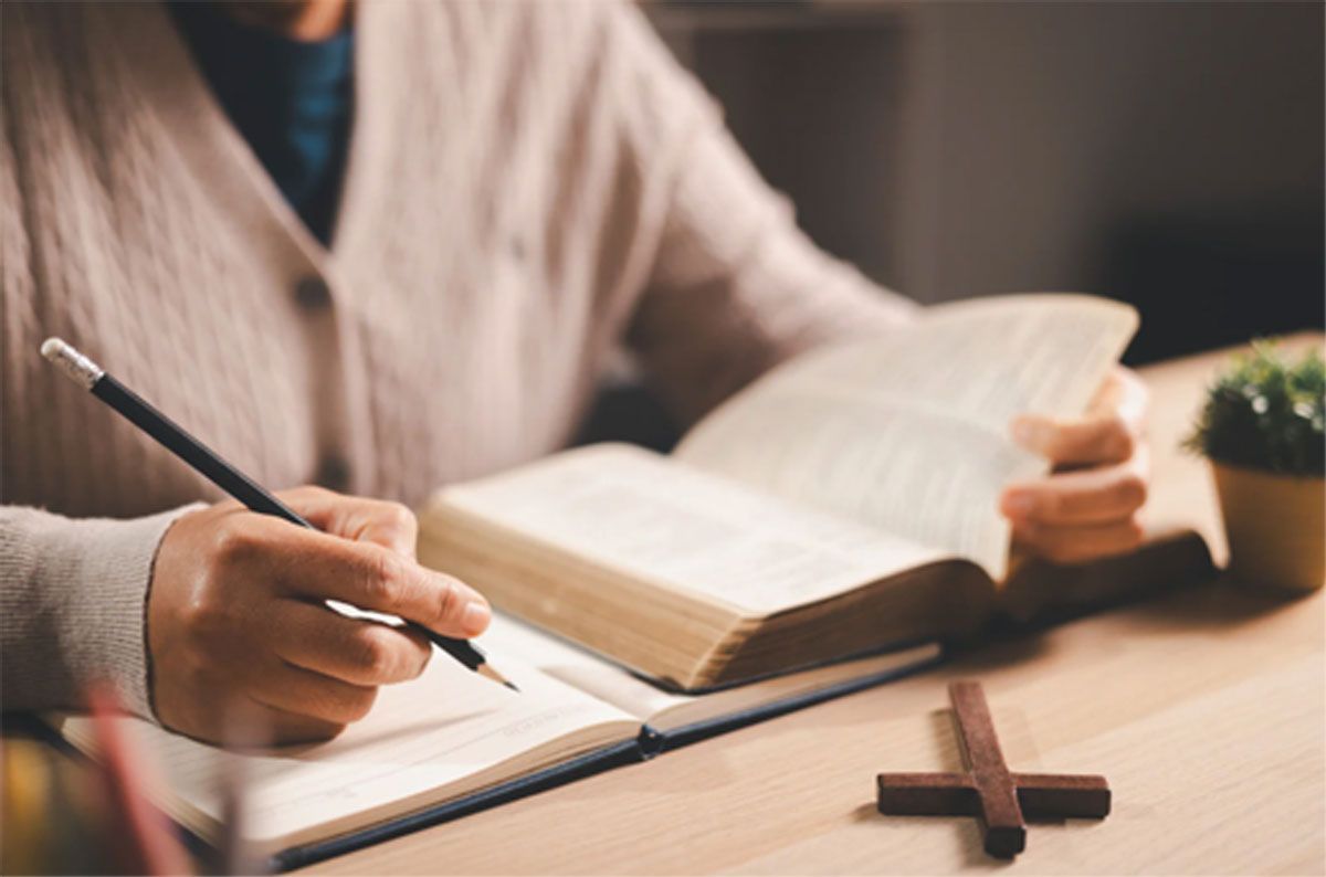Hand writing in a notebook beside an open book and a cross on a wooden table
