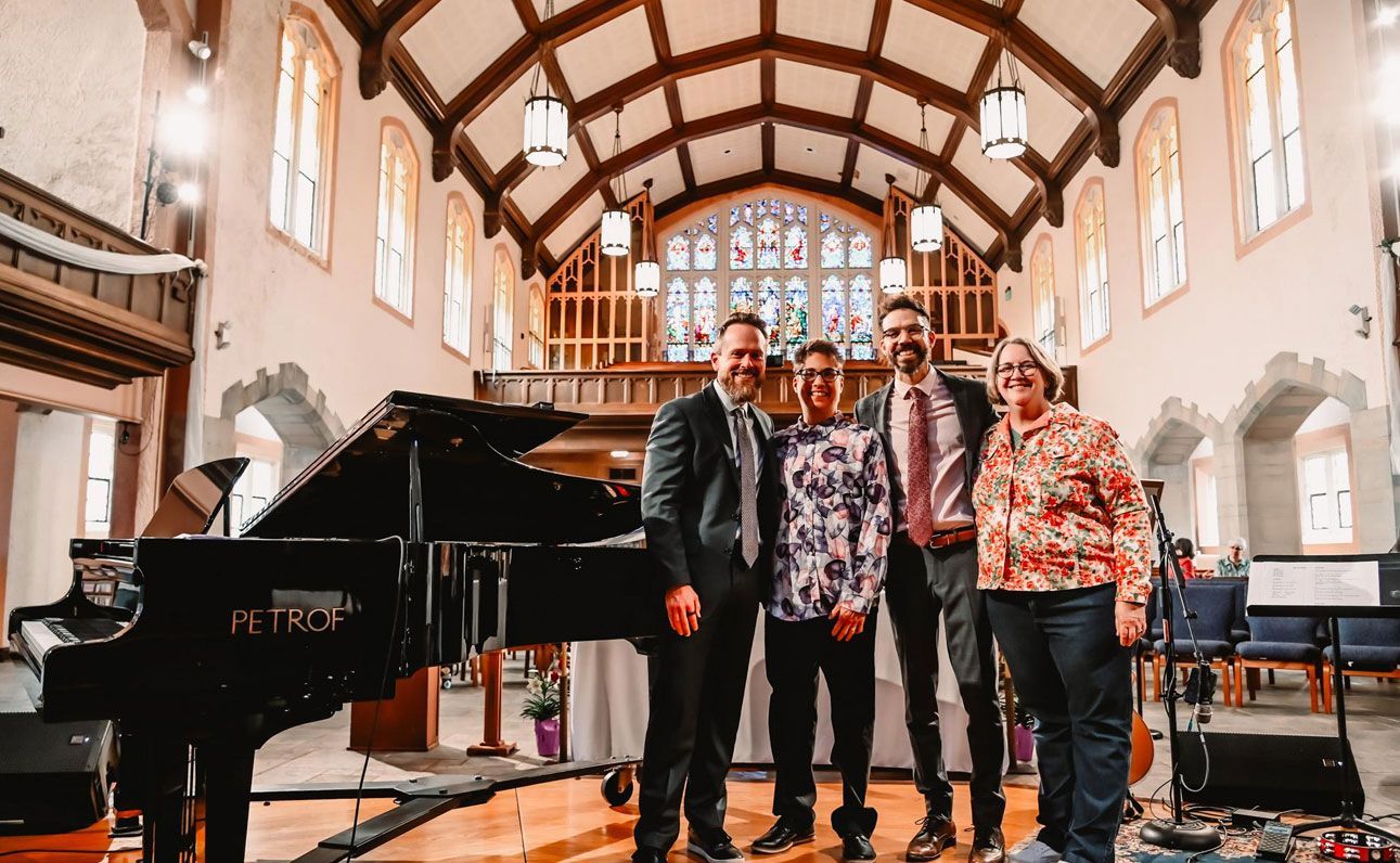 Four musicians pose in a church-like hall beside a grand piano and organ, with stained glass behind them.
