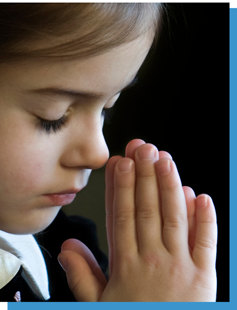 Child with eyes closed, praying with hands pressed together against a dark background.