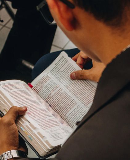 Person reading an open book with small printed text, seated indoors and holding the pages with both hands