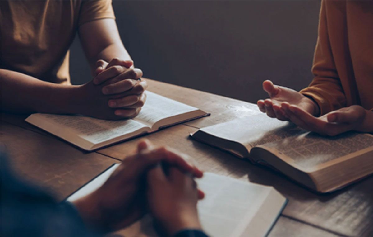 Hands clasped over open books on a wooden table in a dim study group setting