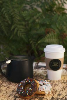 Black mug, white takeaway cup, and two sprinkled donuts on a marble table outdoors.