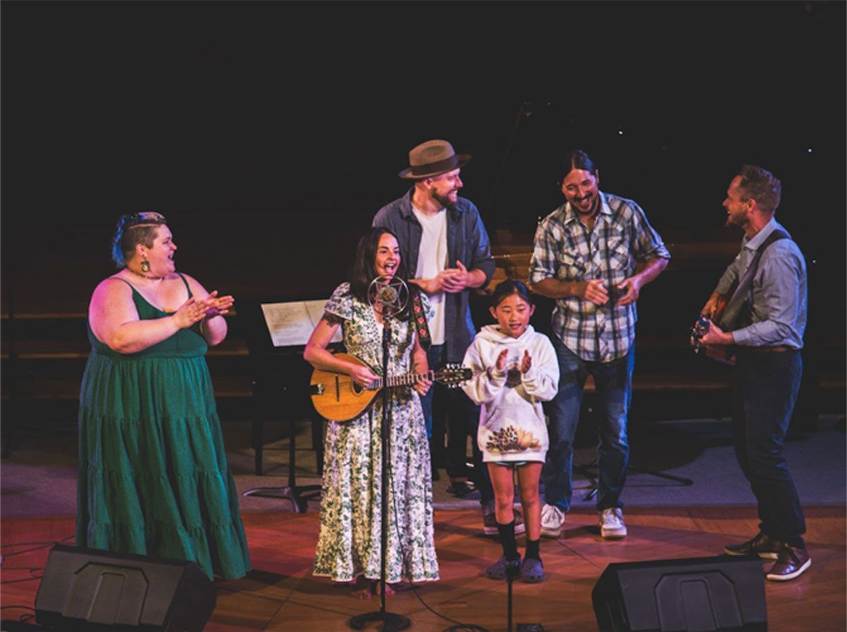 Six musicians on stage, one playing guitar and others singing, with dark background and warm spotlighting
