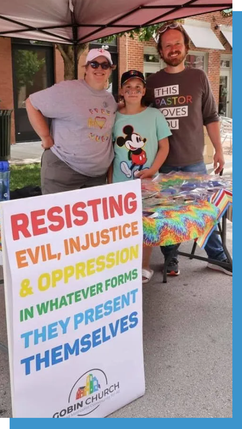 Three people posing behind a sign reading “Resisting evil, injustice & oppression in whatever forms they present themselves”