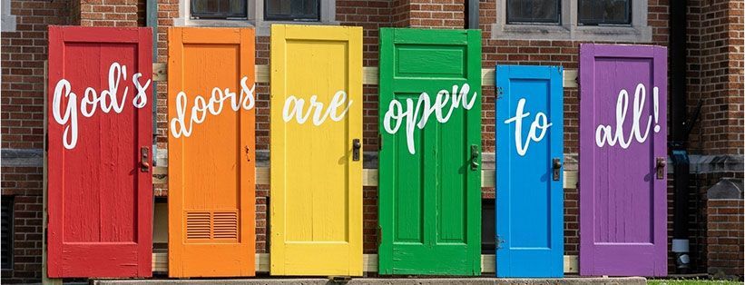Colorful doors spelling “God’s doors are open to all” against a brick wall