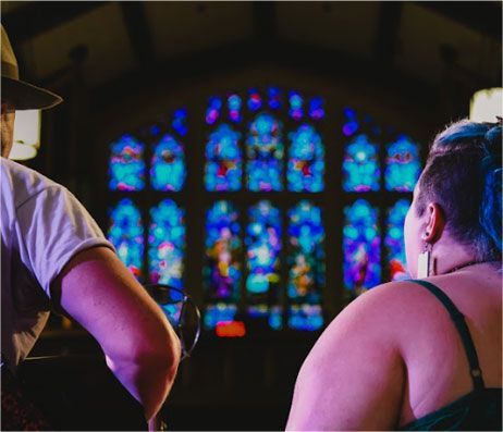 People seated before a colorful stained-glass church window, viewed from behind.