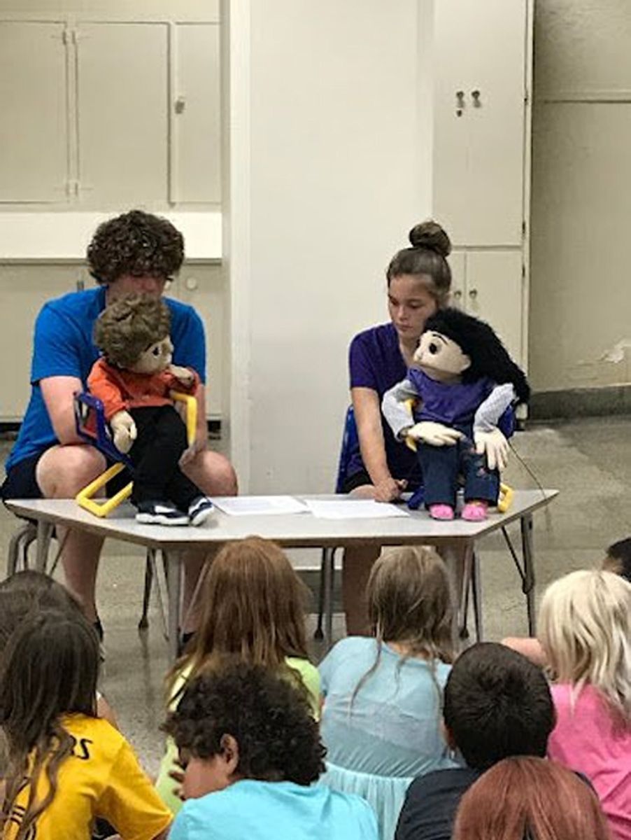 Two puppeteers perform with puppets at a table for an audience of children.