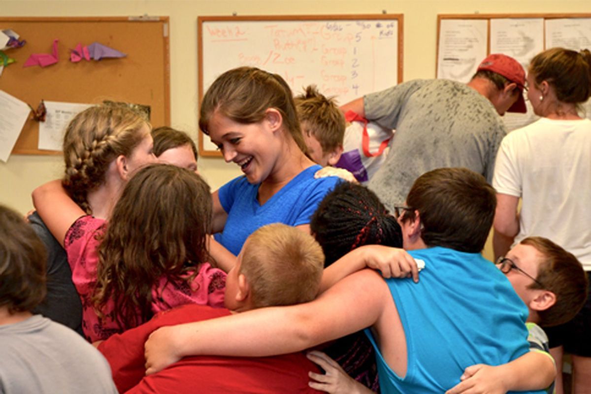 Group hug in a classroom, with smiling people gathered around a woman in a blue shirt.