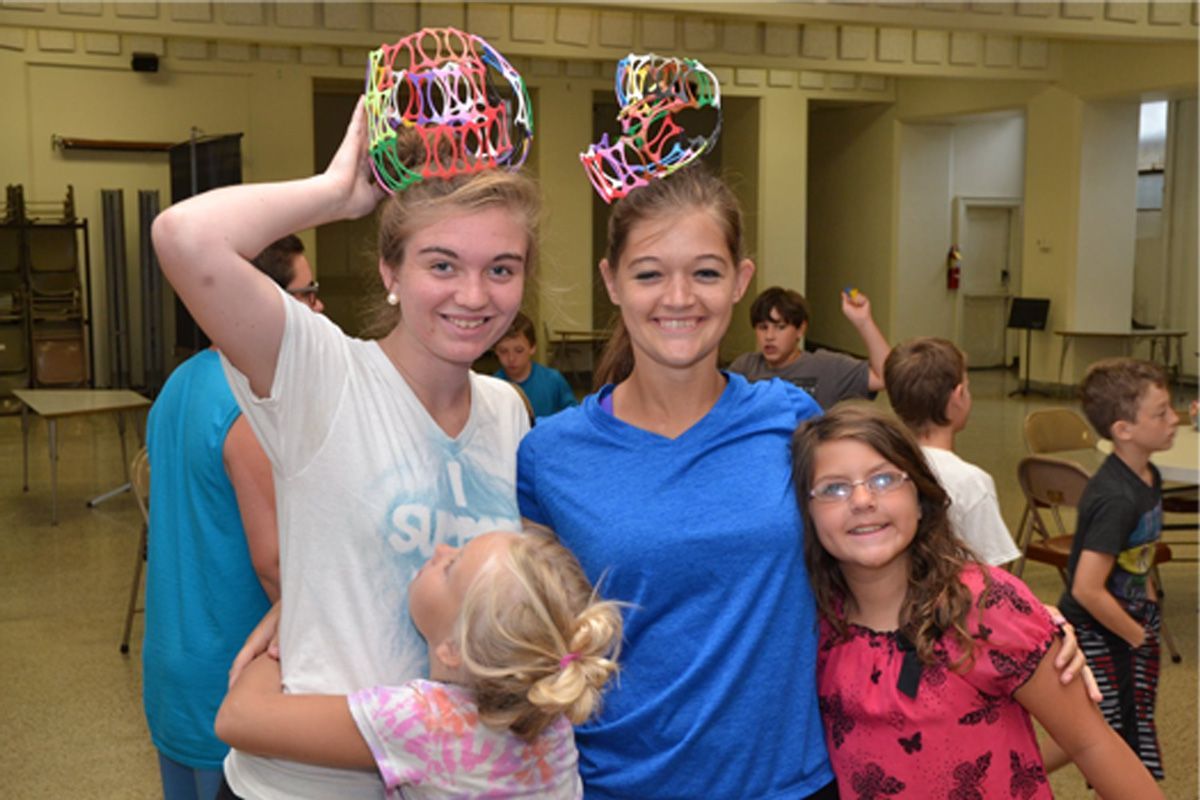 Four people in a hall pose smiling, wearing colorful paper crowns and a pink shirt.