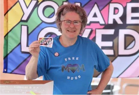 Smiling person in blue shirt holding up a hand sign in front of a colorful “You are loved” banner