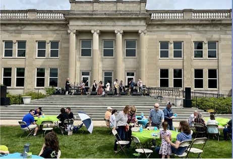 People sitting at tables on a lawn in front of a large stone building with steps and columns
