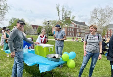 People at a campus outdoor event beside a blue table with green and blue balloons and a food container