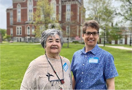 Two people standing on a grassy lawn in front of a brick building, smiling at the camera.