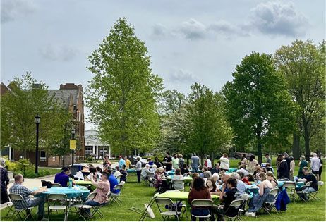 People seated at tables in a sunny park with green trees and grass
