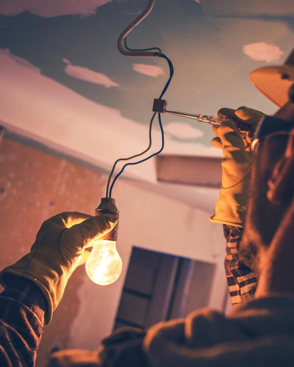Hands wiring a hanging light bulb indoors, with exposed cords and a warm glow.