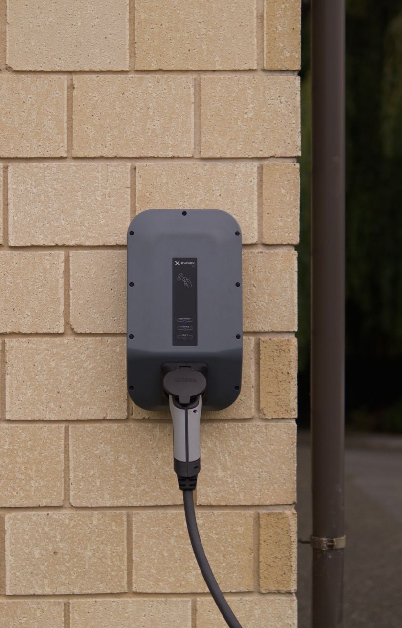 Wall-mounted EV charger with plugged-in cable on a beige brick exterior wall.