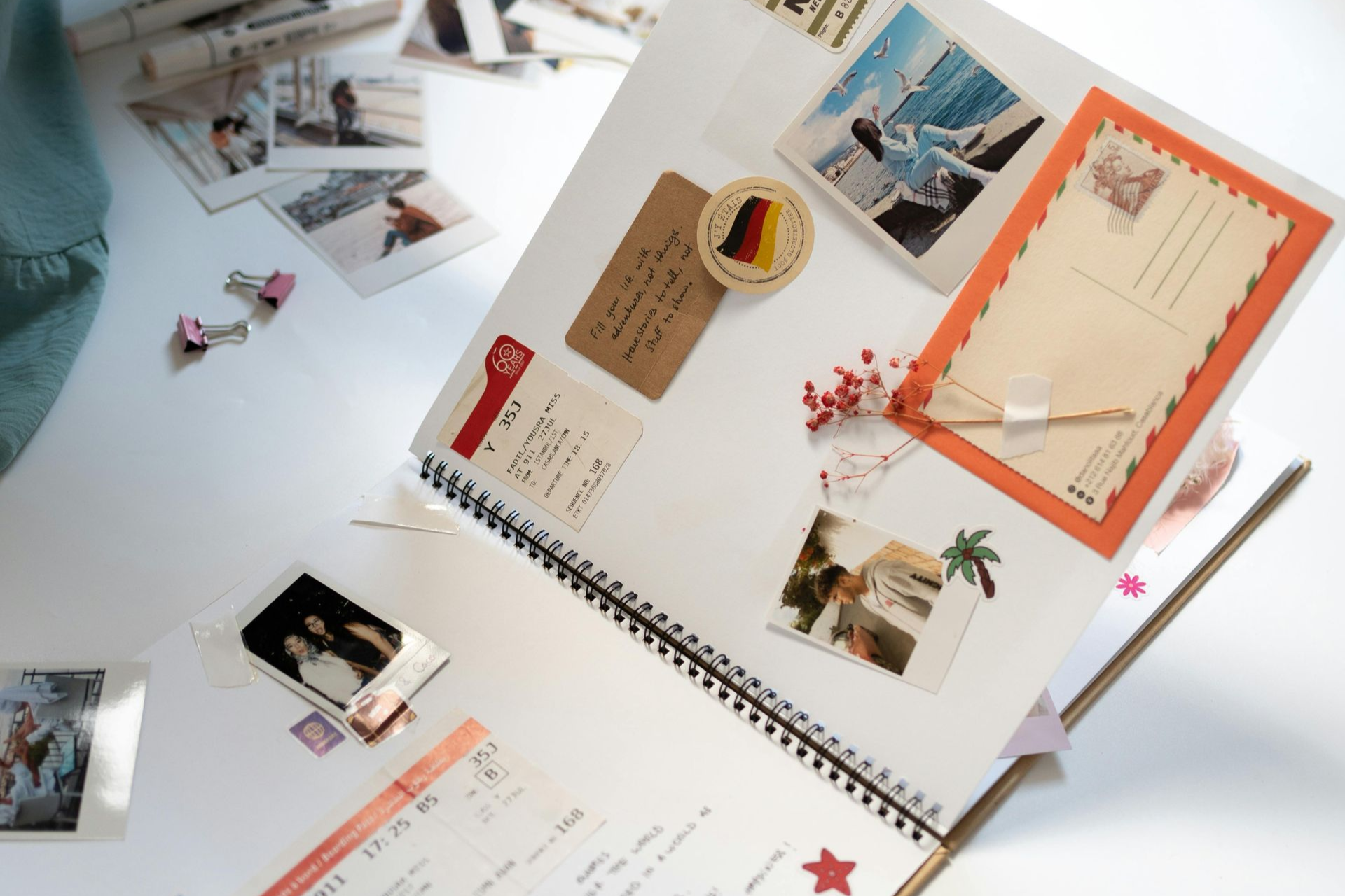 Een open plakboek met foto's, ansichtkaarten en decoratief briefpapier op een wit bureau met schrijfwaren in de buurt.