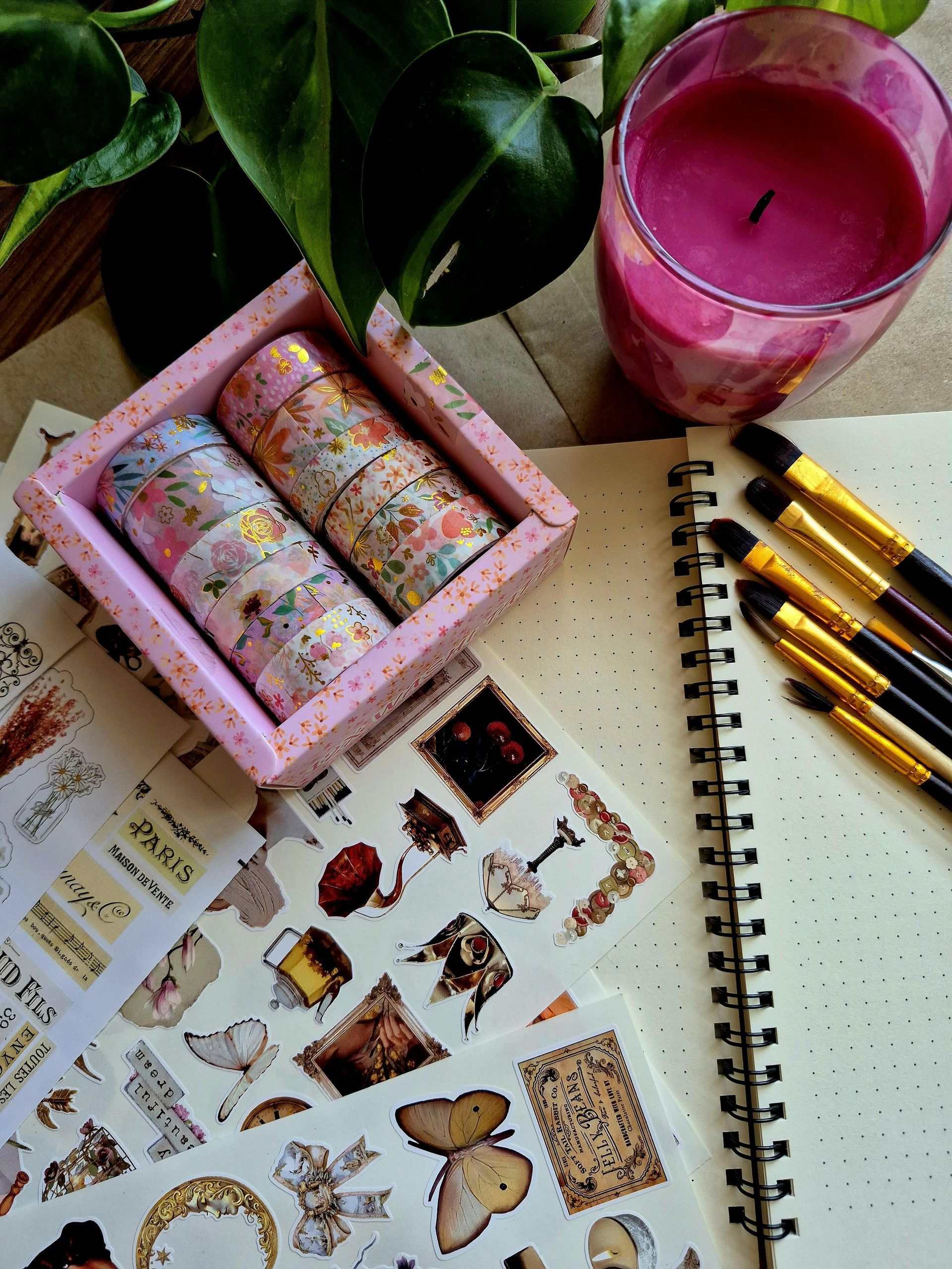 Rollen washi-tape, stickervellen en penselen liggen uitgestald op een bureau naast een potplant en een roze kaars.