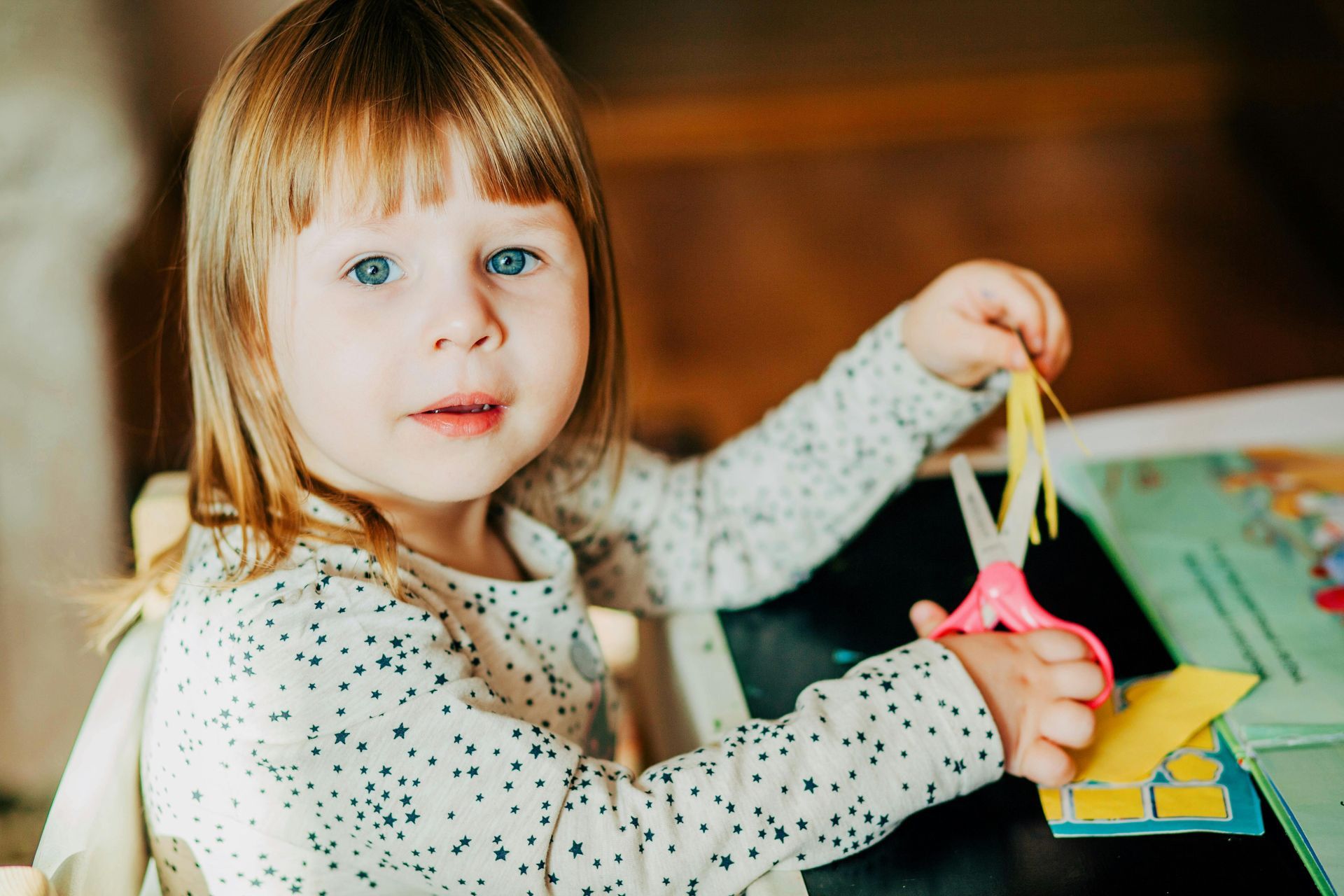 Een kind met blond haar en een topje met print gebruikt een roze schaar om een ​​gele strook papier aan een tafel te knippen.
