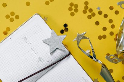 Een notitieboekje met open raster, een zilveren ster en een zilveren taartschep op een felgele achtergrond met confetti.