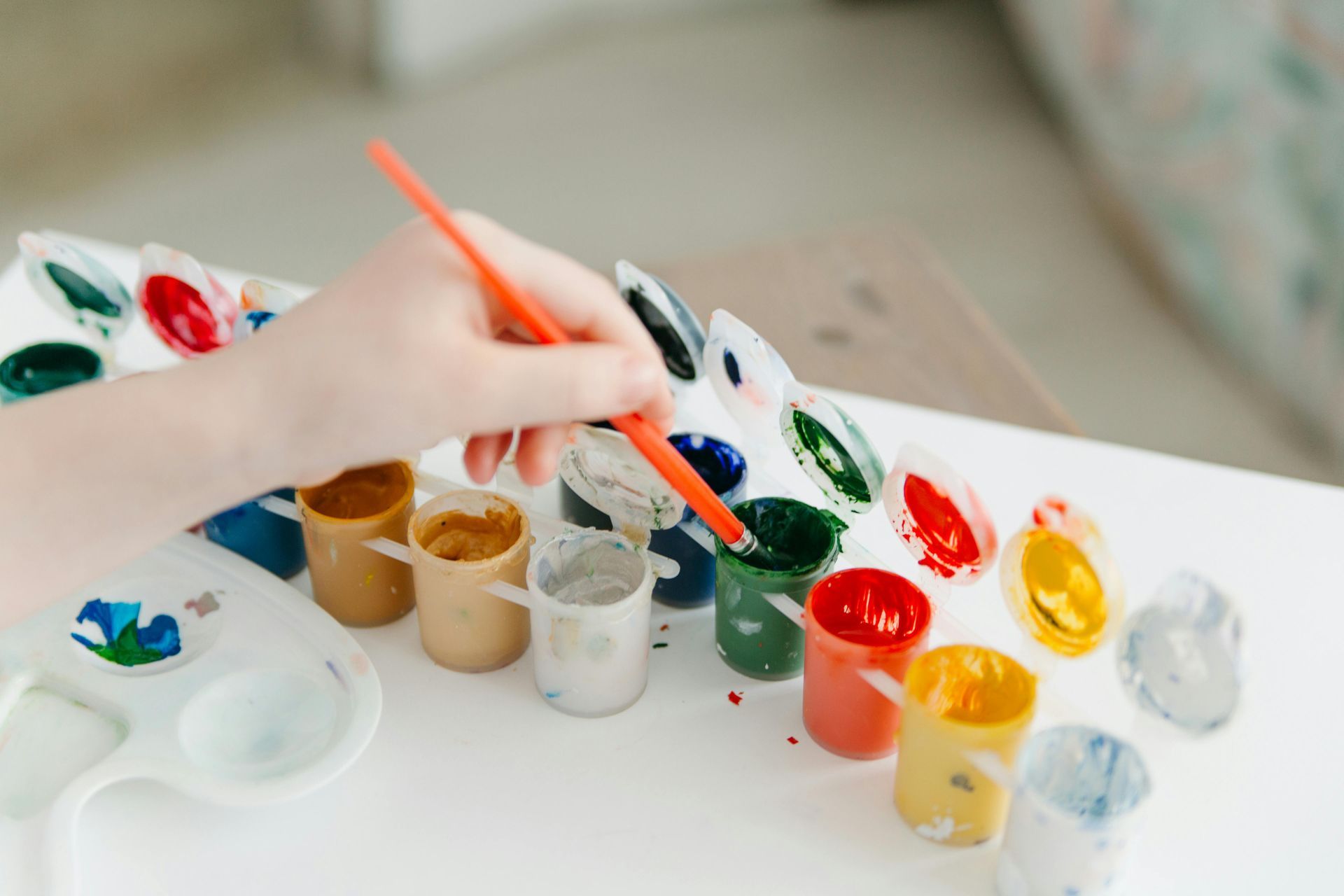 Een hand gebruikt een oranje kwast om in een klein bakje donkergroene verf te dippen, dat deel uitmaakt van een set open potjes acrylverf.