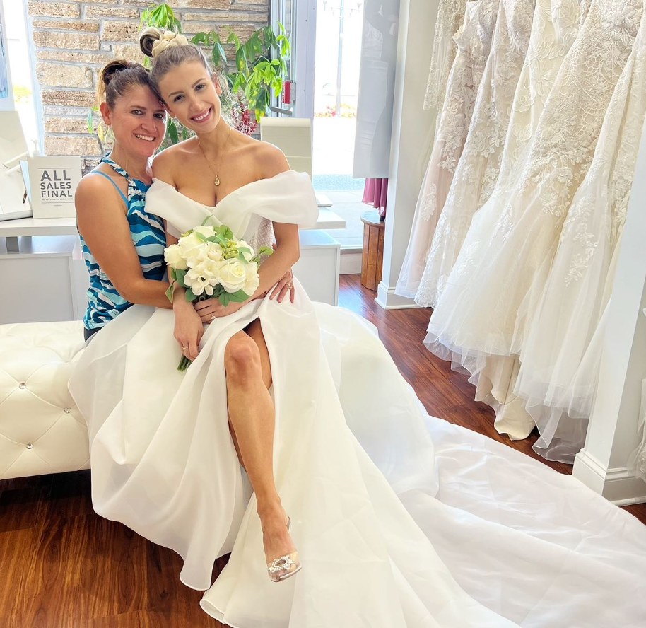 Woman in wedding dress, friend, holding flowers, smiling, sitting in bridal shop.