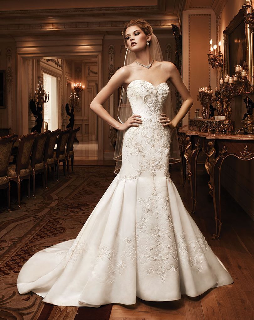Woman in white strapless mermaid-style wedding gown poses indoors with hands on hips, gazing upwards.