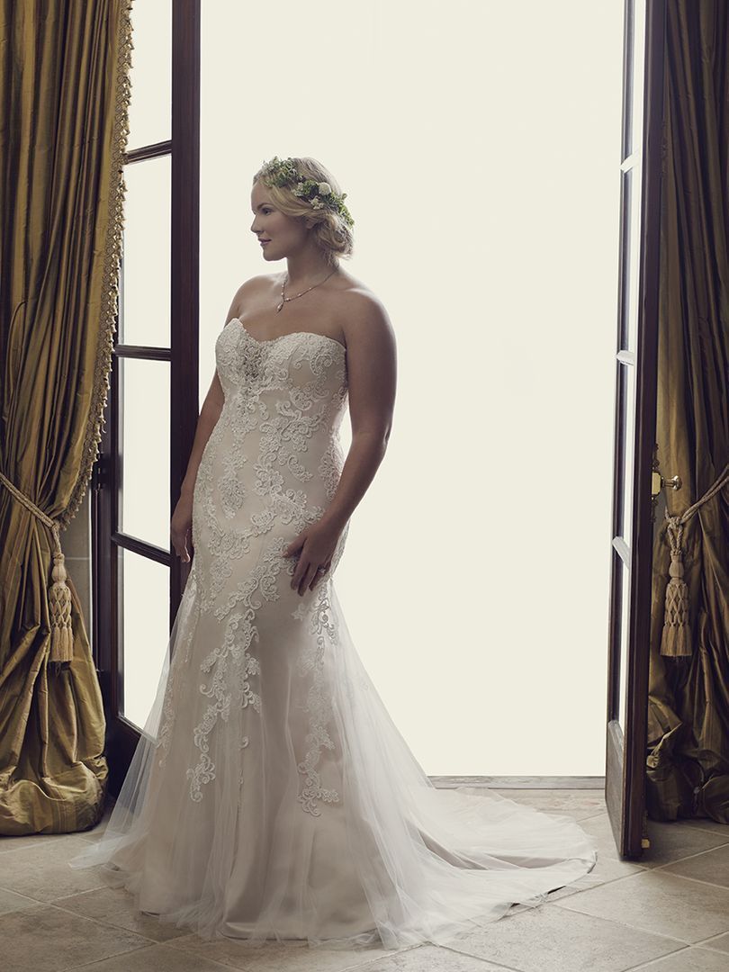 Woman in a white wedding dress stands by open doors with gold curtains; she looks to the side with a smile.